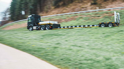 Forsttiefbett Sattelauflieger, der auf einem grasbewachsenen Straßenrand fährt und für den Transport von großen und schweren Forstmaschinen bestimmt ist.